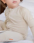 A baby sitting on a bed wearing the **Ramly Pyjama** from the **Grow with Nohm: Gift Set**, a beige bamboo one-piece featuring a front zipper for easy changes and a snug, comfy fit.