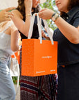 A person holding the **Neutral Nohm Gift Box** in its signature bright orange packaging with white handles, surrounded by others at an outdoor event.