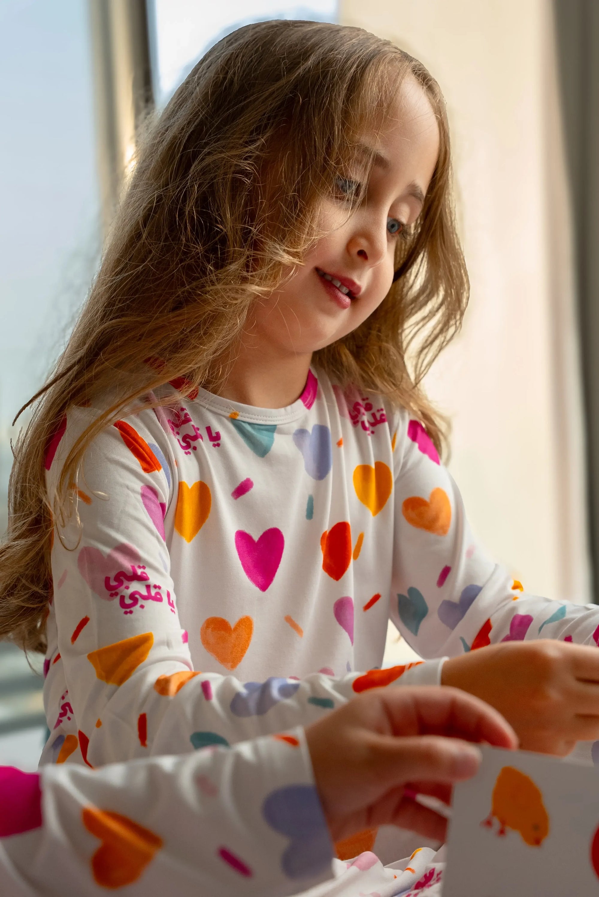 A girl wearing our Albi Long Sleeve Pyjama, smiling softly as she plays and explores colorful cards.
