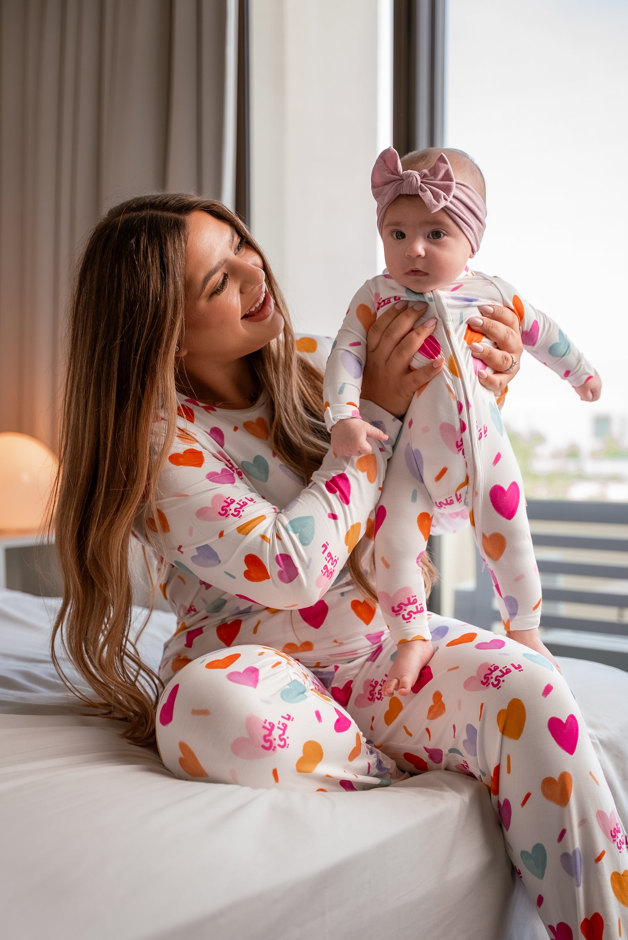 Mother and baby wearing matching Albi Pyjamas, sharing a joyful moment on the bed surrounded by colorful heart prints and cozy morning light.