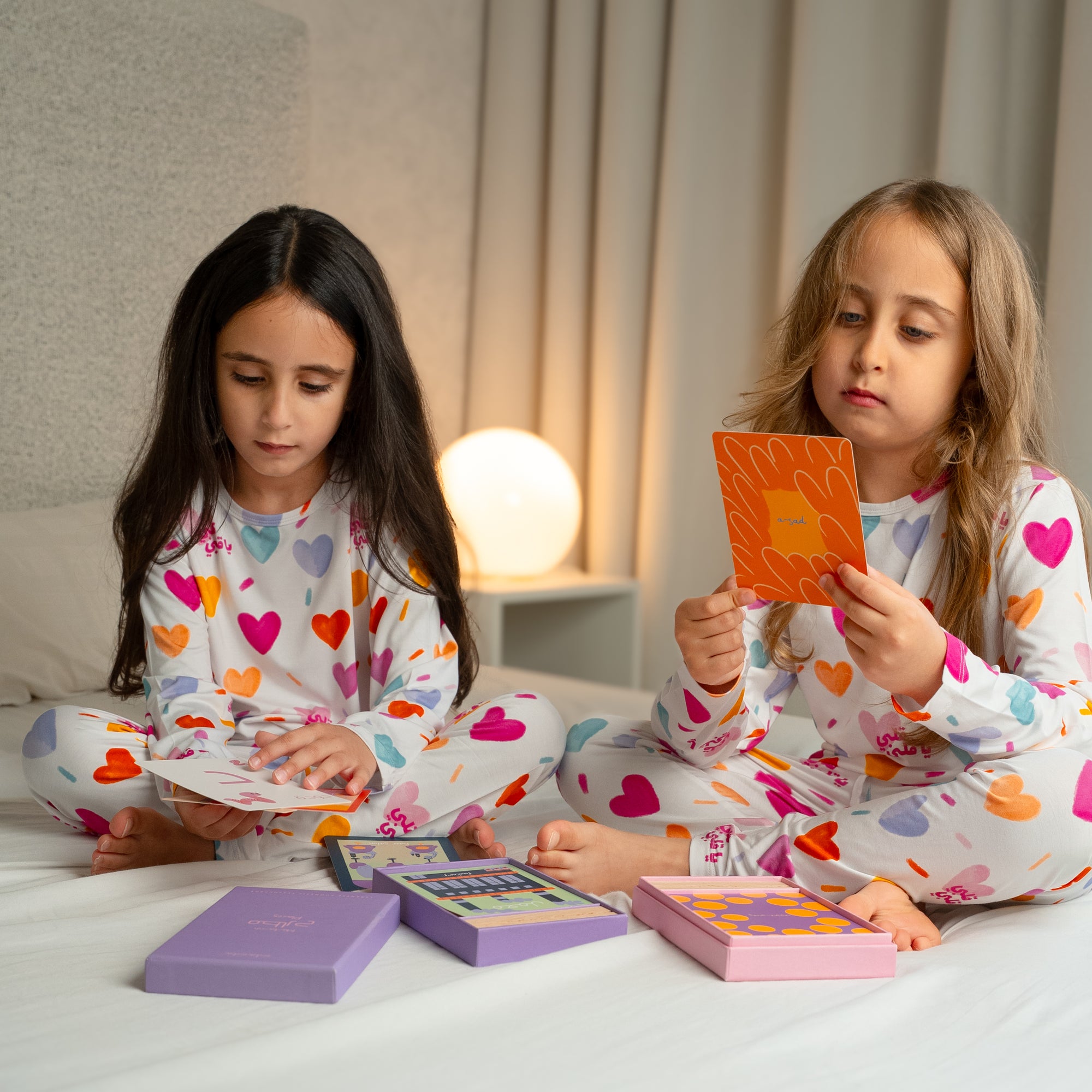 Two girls wearing our Albi Long Sleeve Pyjama, enjoying a cozy playtime together with colorful cards on the bed.