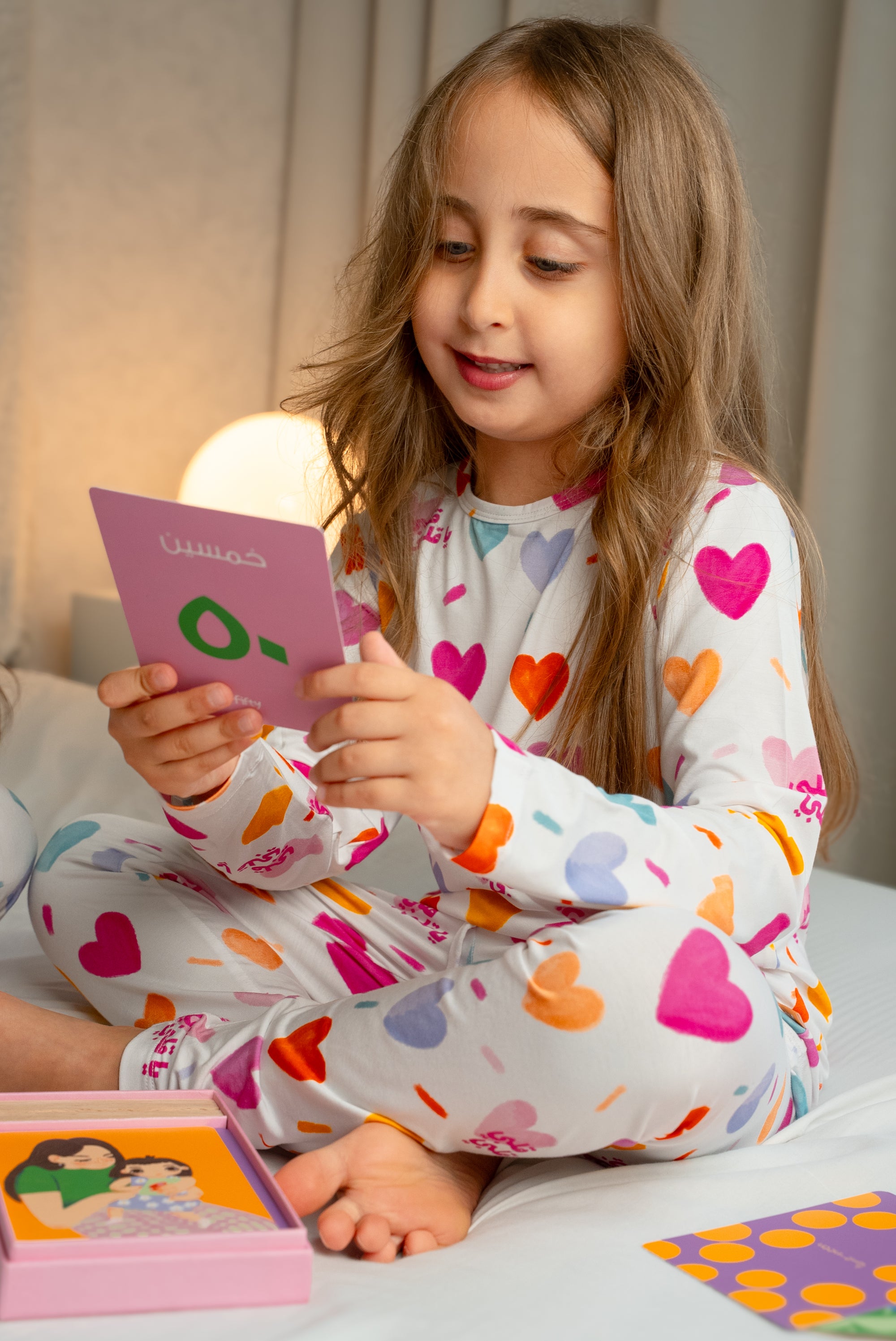 A girl wearing our Albi Long Sleeve Pyjama, smiling as she reads a colorful card during playtime.
