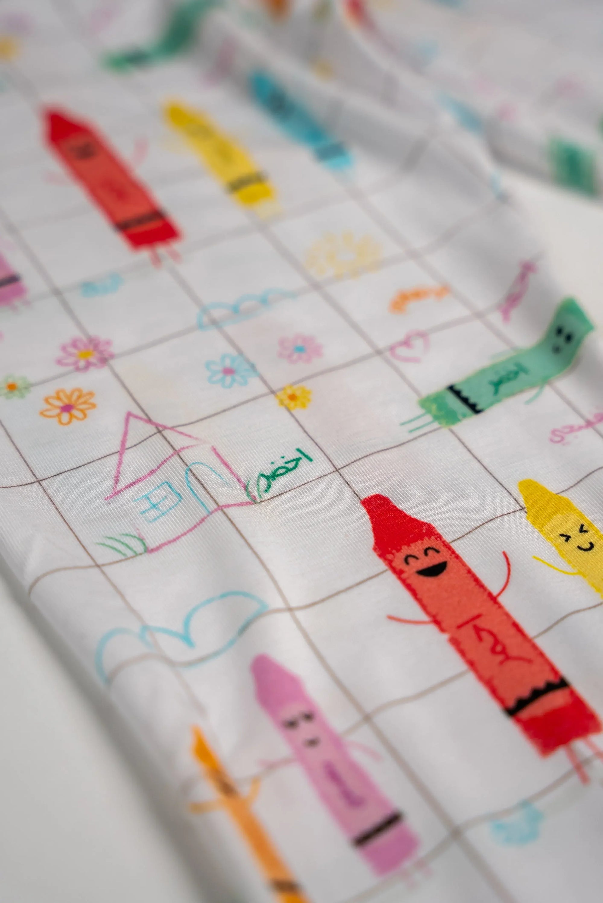 Close-up of our Crayons Pyjama fabric, featuring colorful Arabic crayon characters and playful hand-drawn doodles on a soft grid pattern.