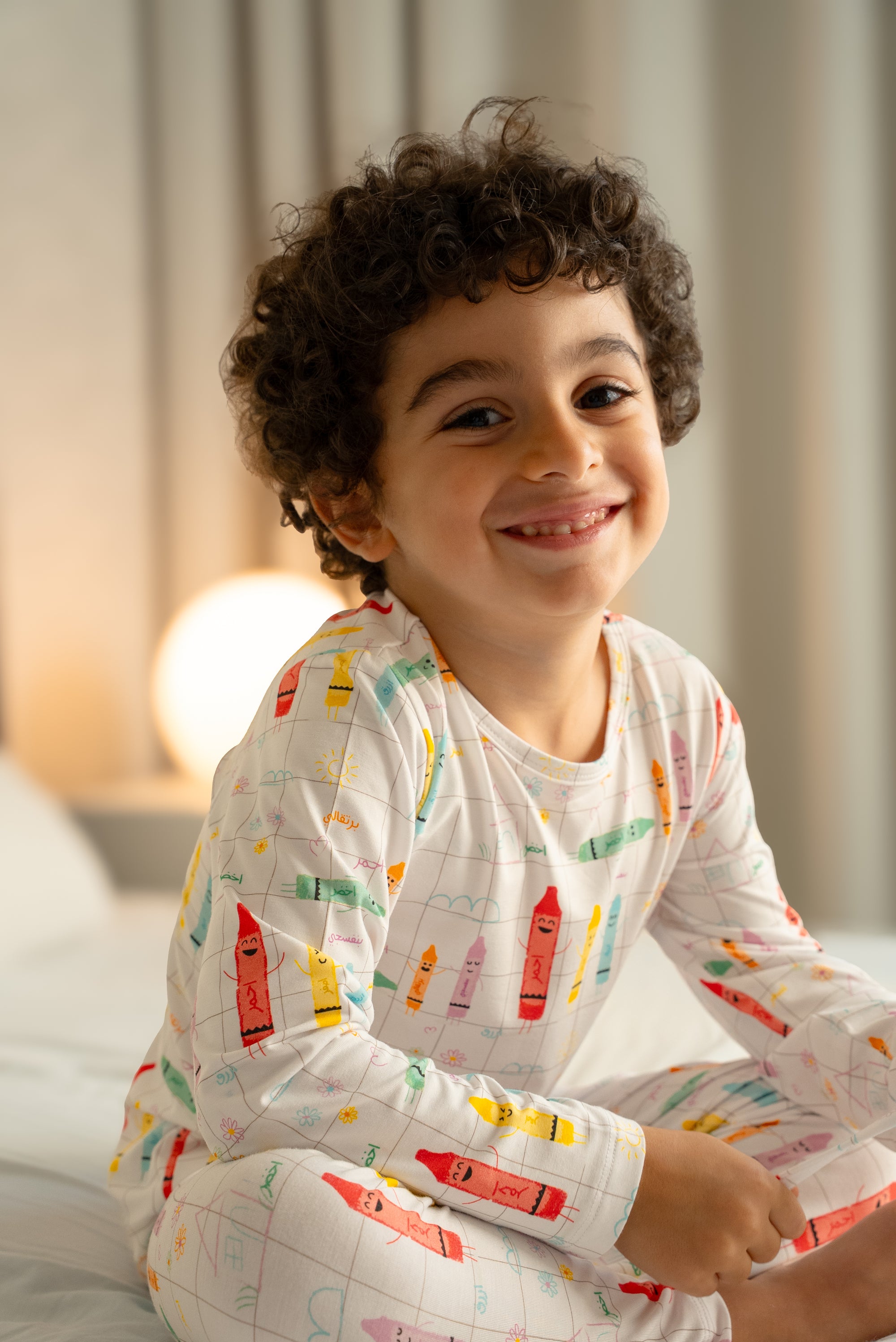 Child wearing colourful our Crayons Pyjama, smiling while sitting on a bed.