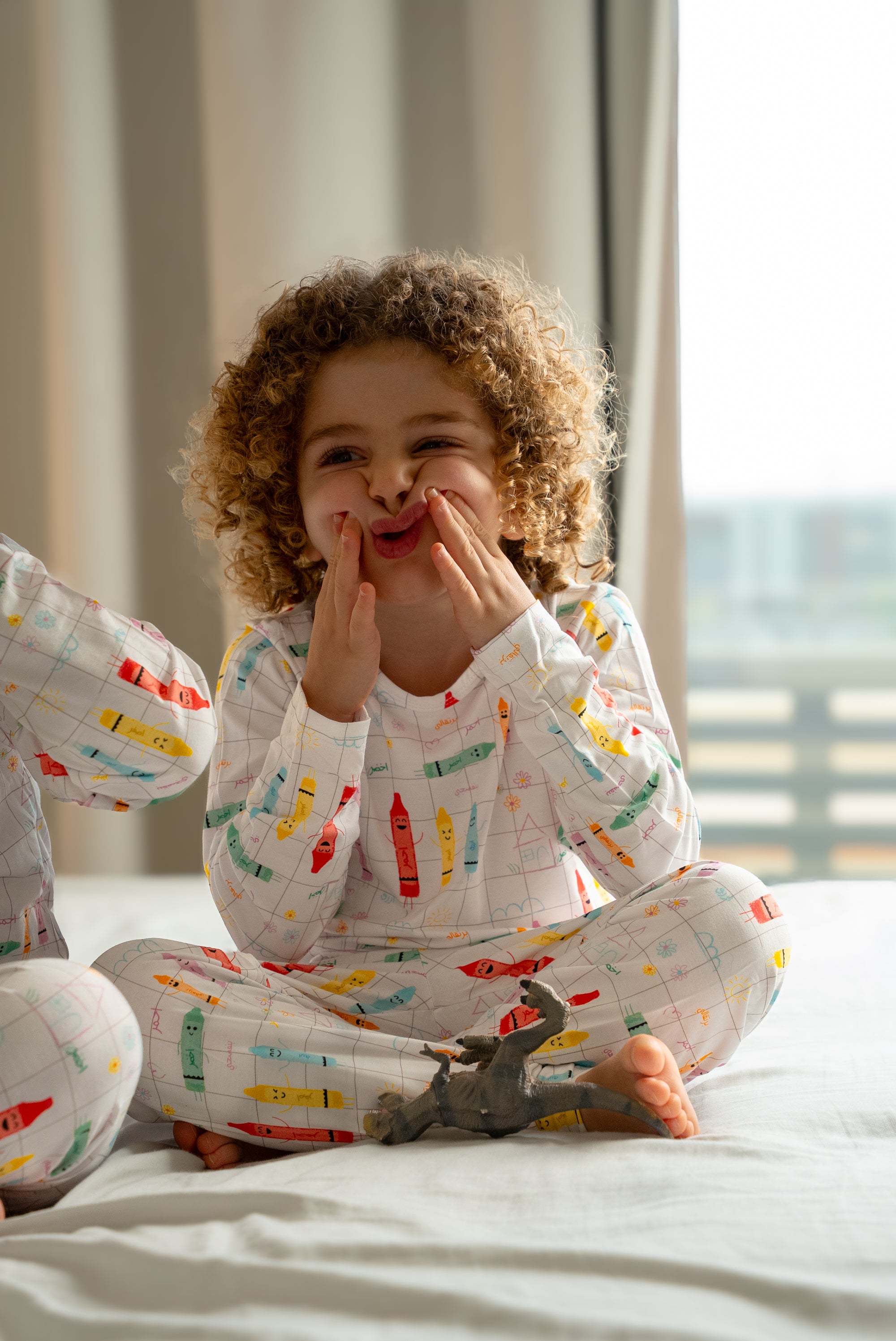 Child wearing our Crayons Pyjama, sitting cross-legged on a bed and playfully making a funny face with her hands.