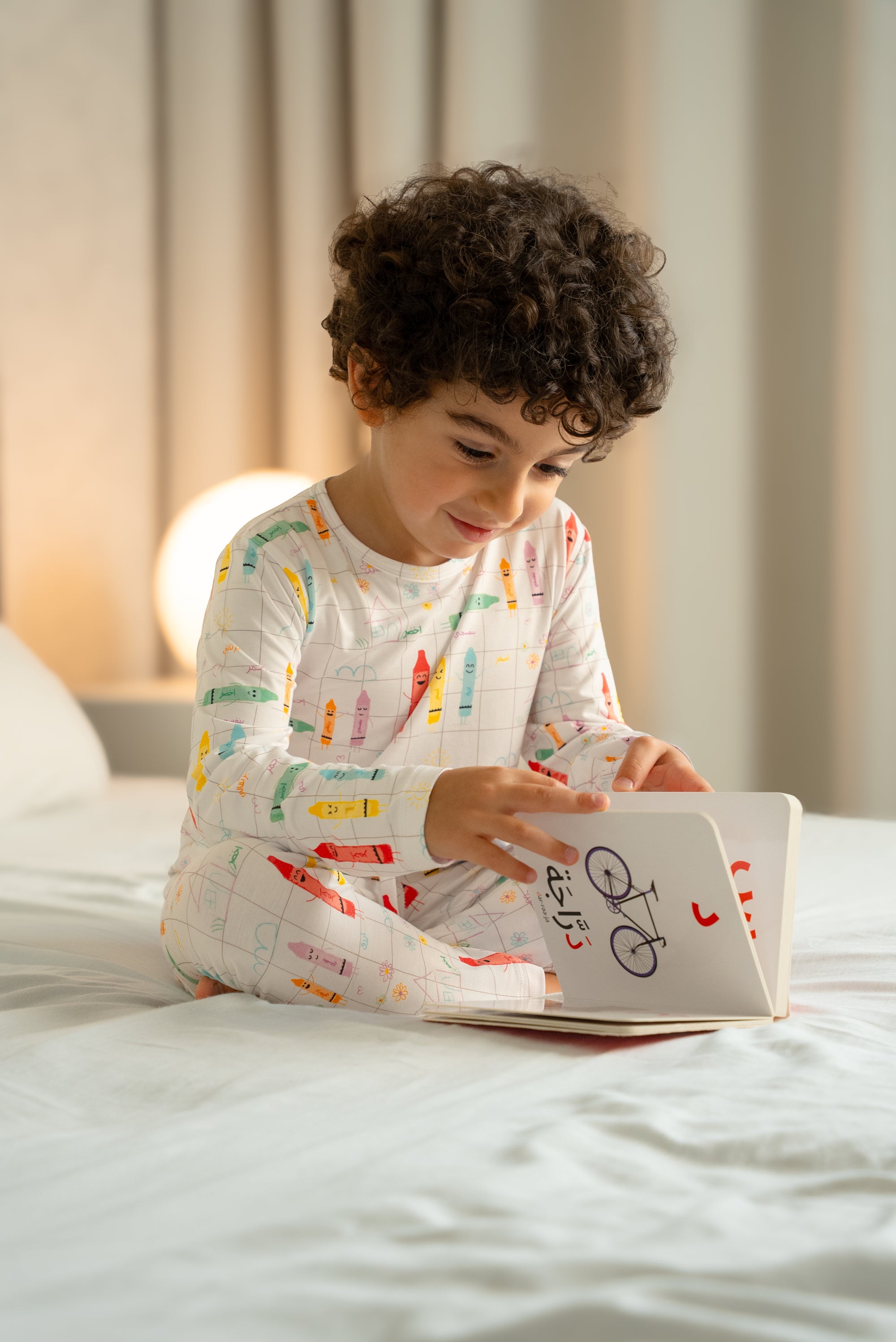 Child wearing our Crayons Pyjama, sitting on a bed and reading an Arabic picture book, surrounded by a warm bedtime glow.