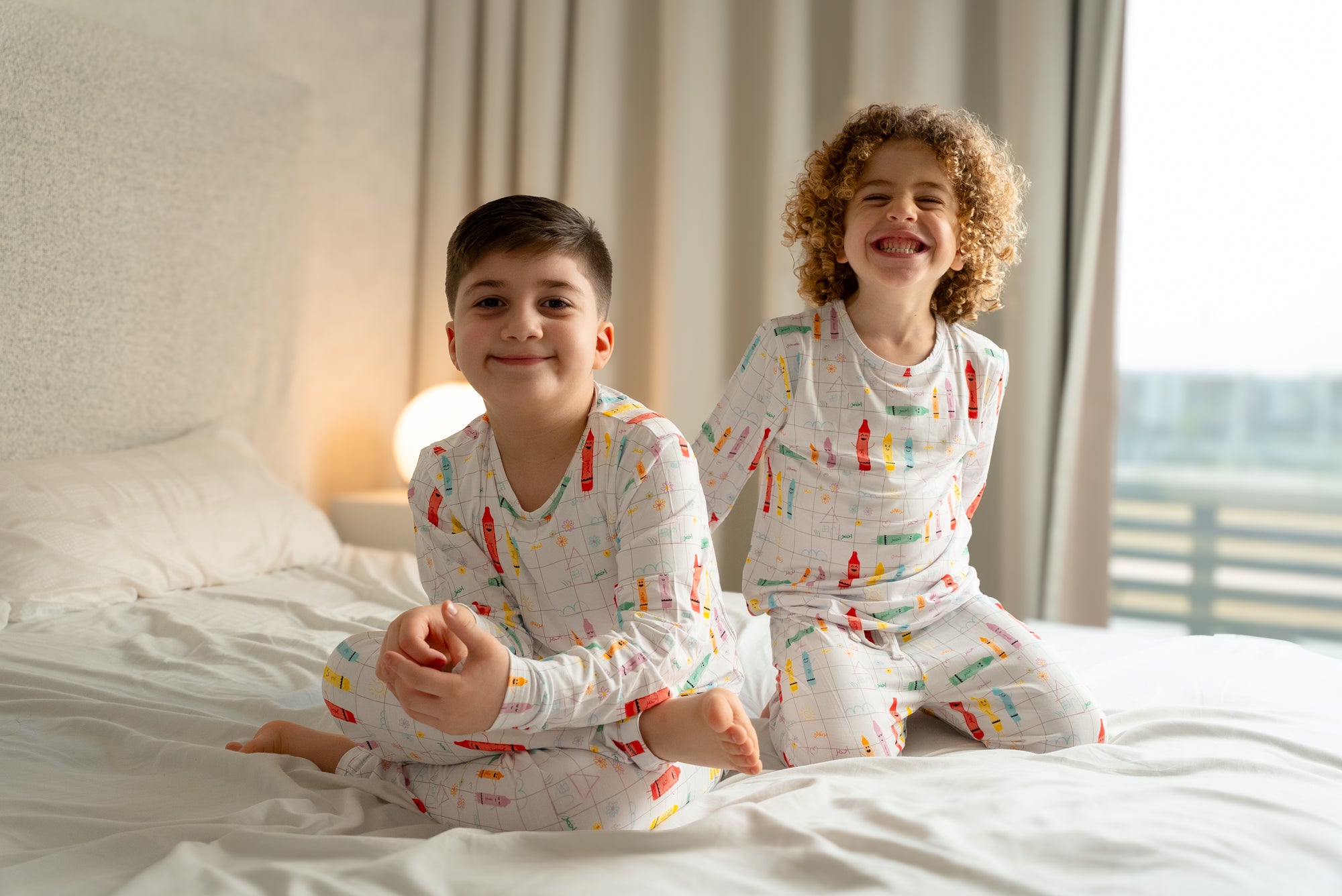 Two children wearing our Crayons Pyjama, sitting and smiling on a bed, enjoying a bright and playful morning together.