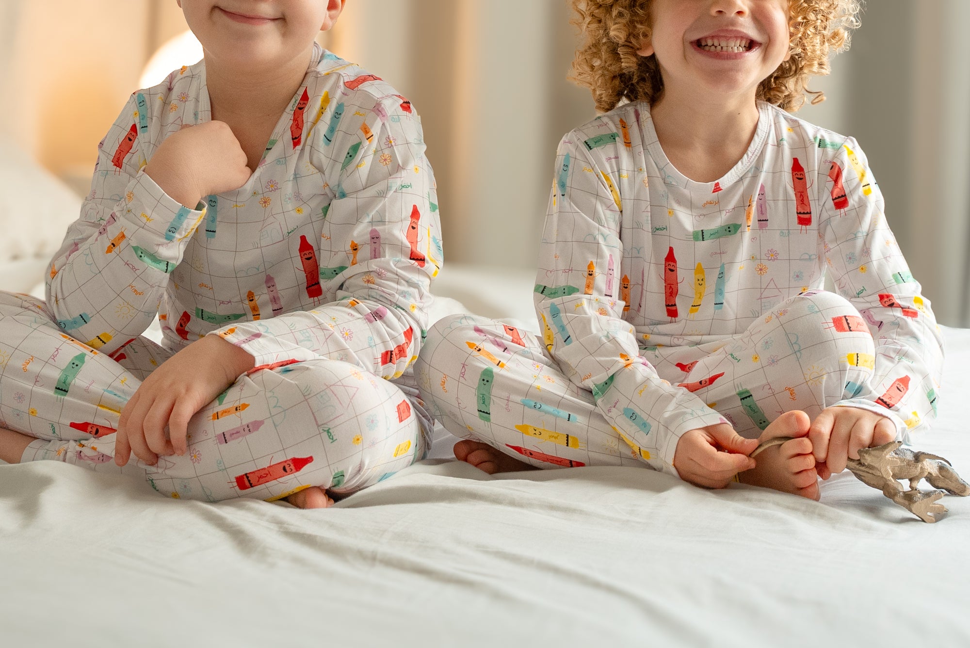 Close-up of two children sitting on a bed, smiling and wearing our Crayons Pyjama with playful Arabic crayon prints.
