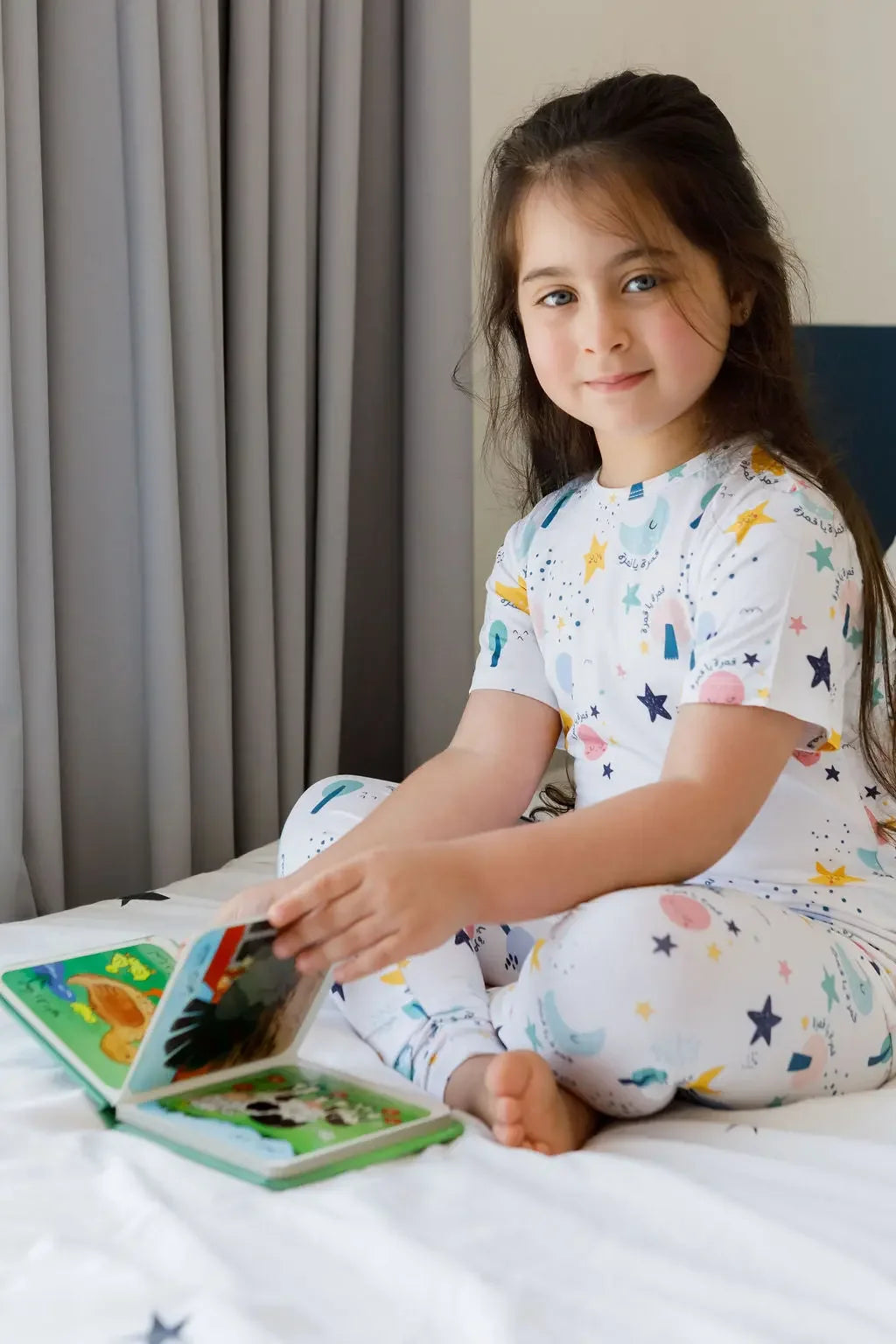 A girl wearing our Amara Short Sleeve Pyjama, enjoying story time in dreamy moon and star prints inspired by “Amara ya Amara.”
