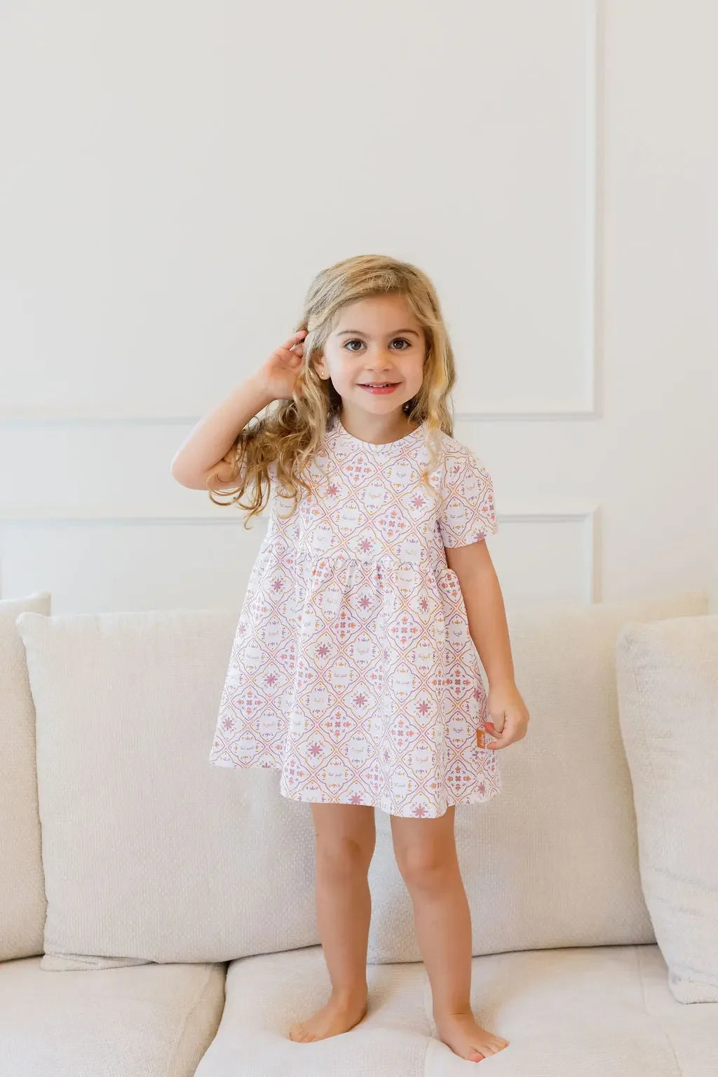 A smiling girl standing on the couch while wearing our Arabesque Dress, showcasing elegant patterns inspired by timeless design.