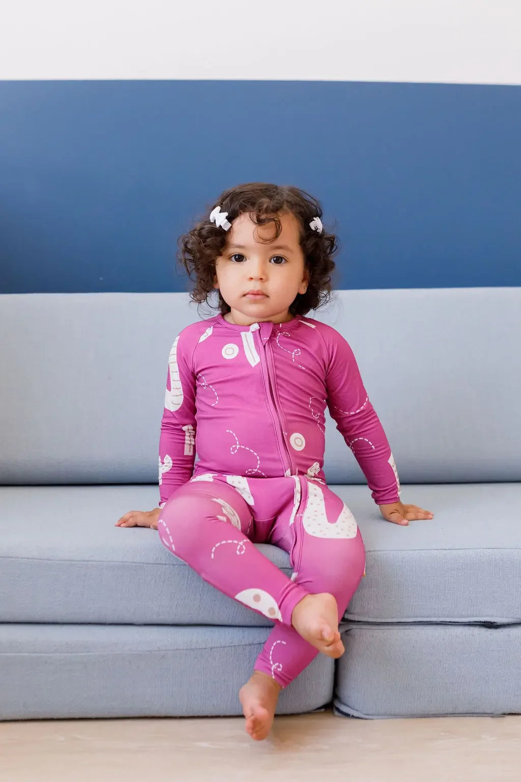 A calm little girl sitting on the couch while wearing our Arqam Smart Baby Pyjama, featuring playful Arabic numbers in a fun magenta design.