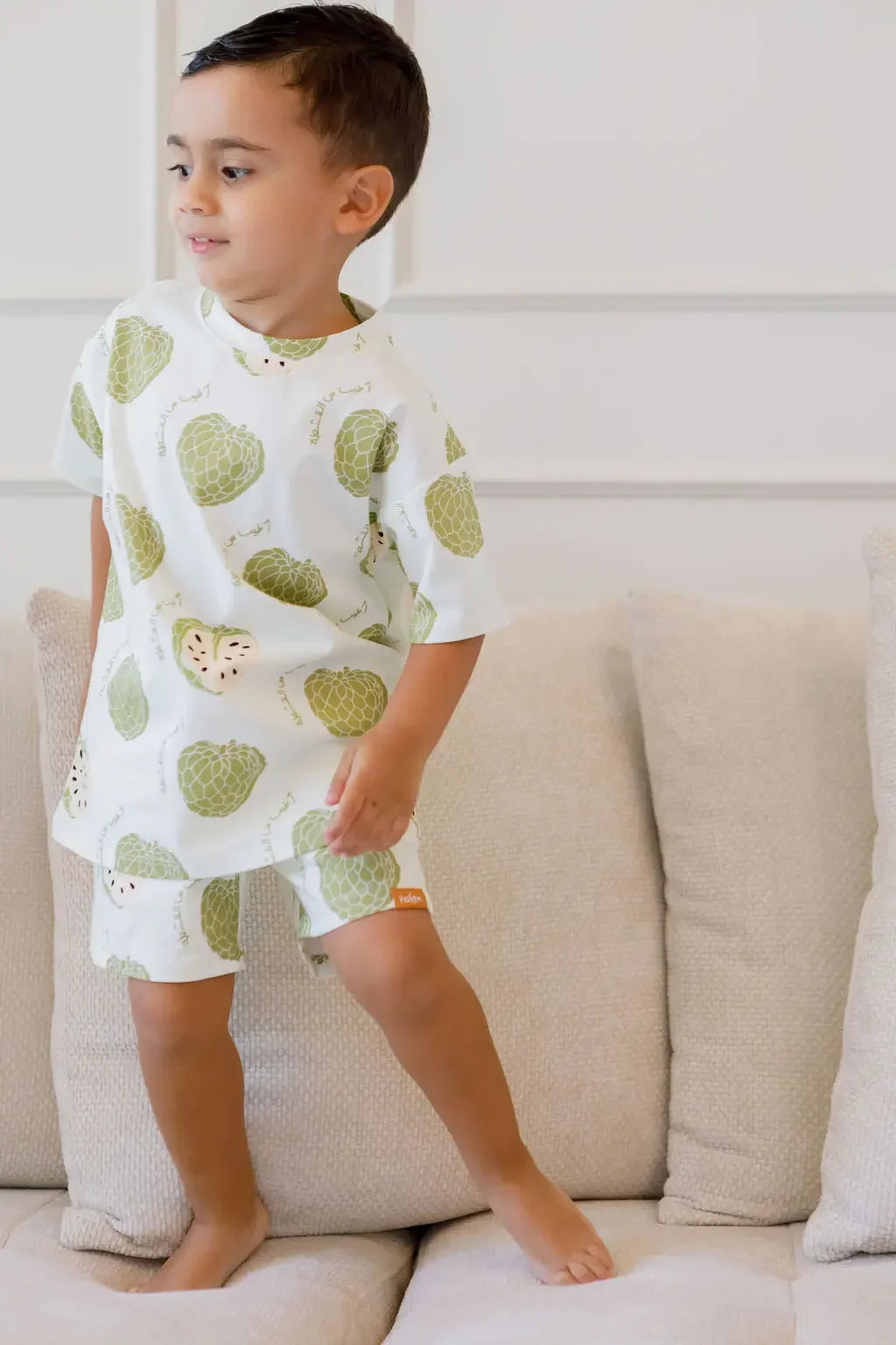 A playful boy standing on the couch while wearing our Ashta Loungewear set, featuring the fun annona fruit design.

