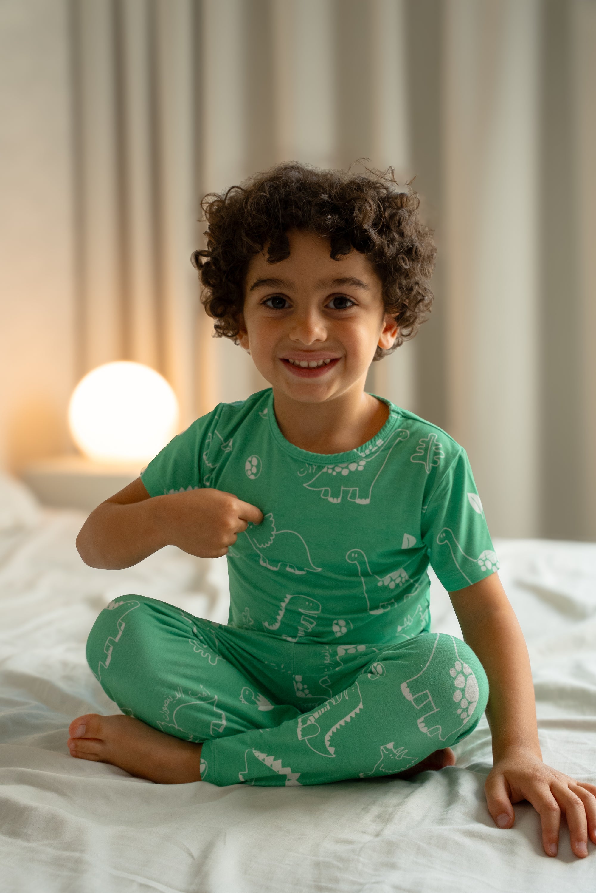 Child wearing our Dinosaur Pyjama, sitting cross-legged on a bed and smiling, showcasing the playful white dinosaur prints on a soft green fabric.