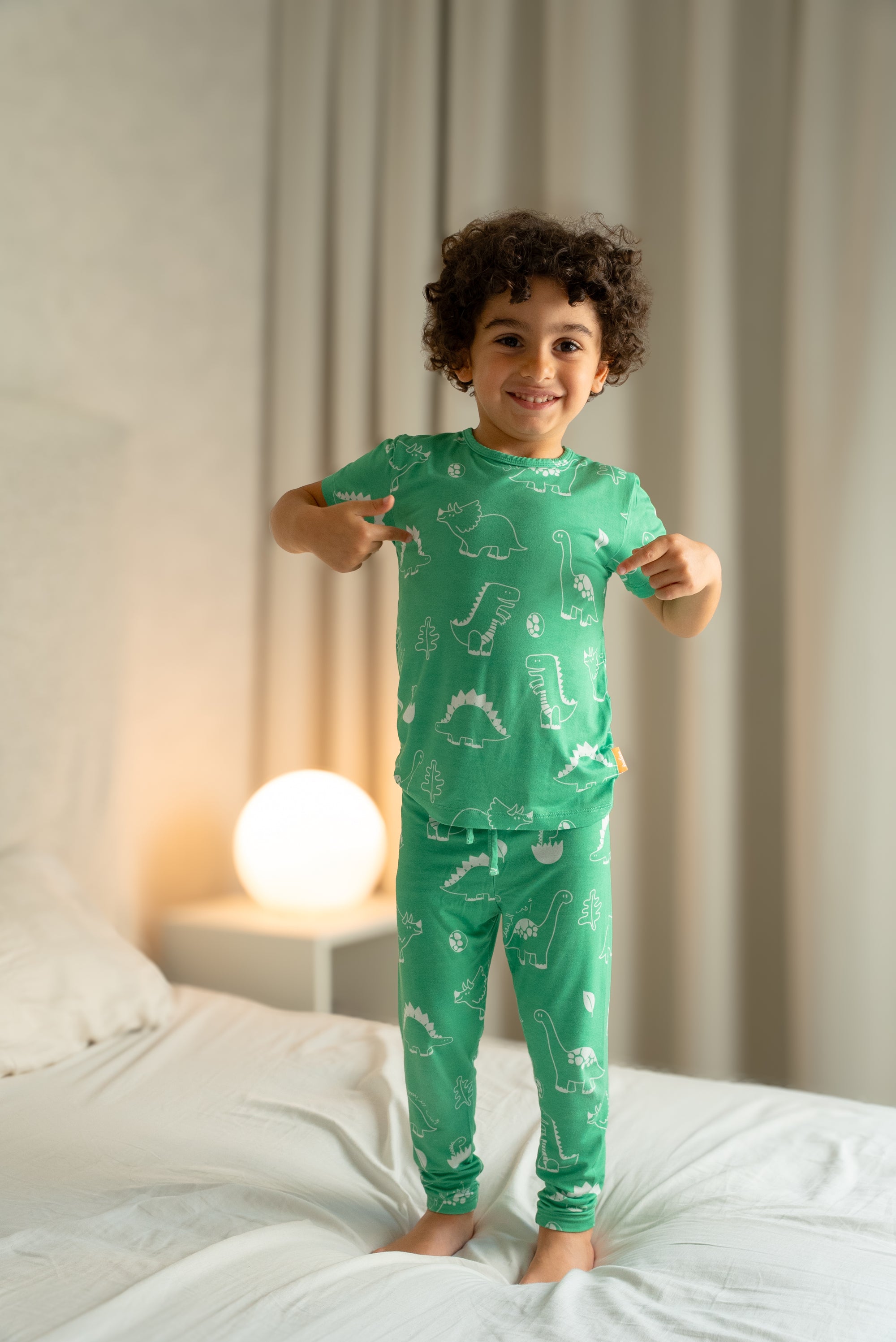 Child wearing our Dinosaur Pyjama, standing on a bed and proudly pointing at the playful white dinosaur prints on the soft green fabric.