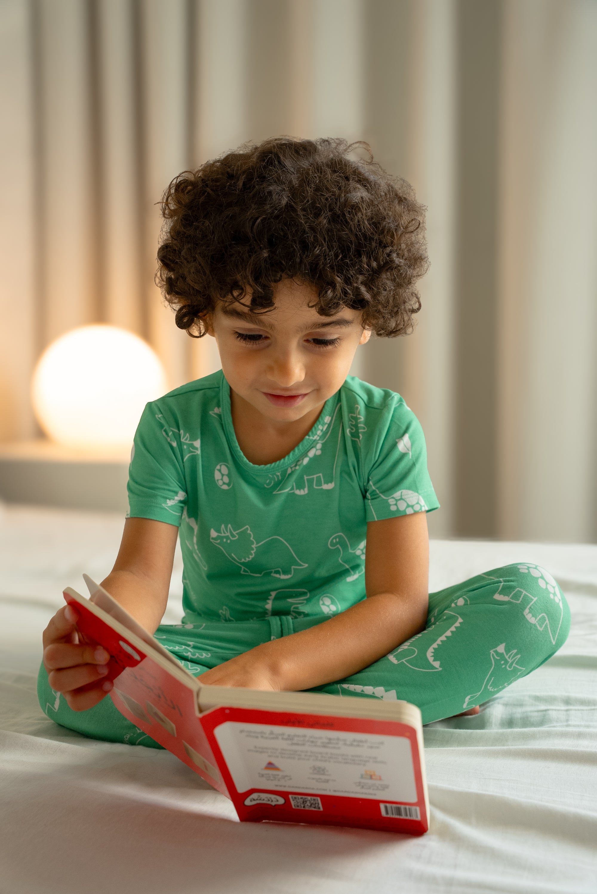 Child wearing our Dinosaur Pyjama, sitting cross-legged on a bed and reading a book, surrounded by a warm bedtime glow.