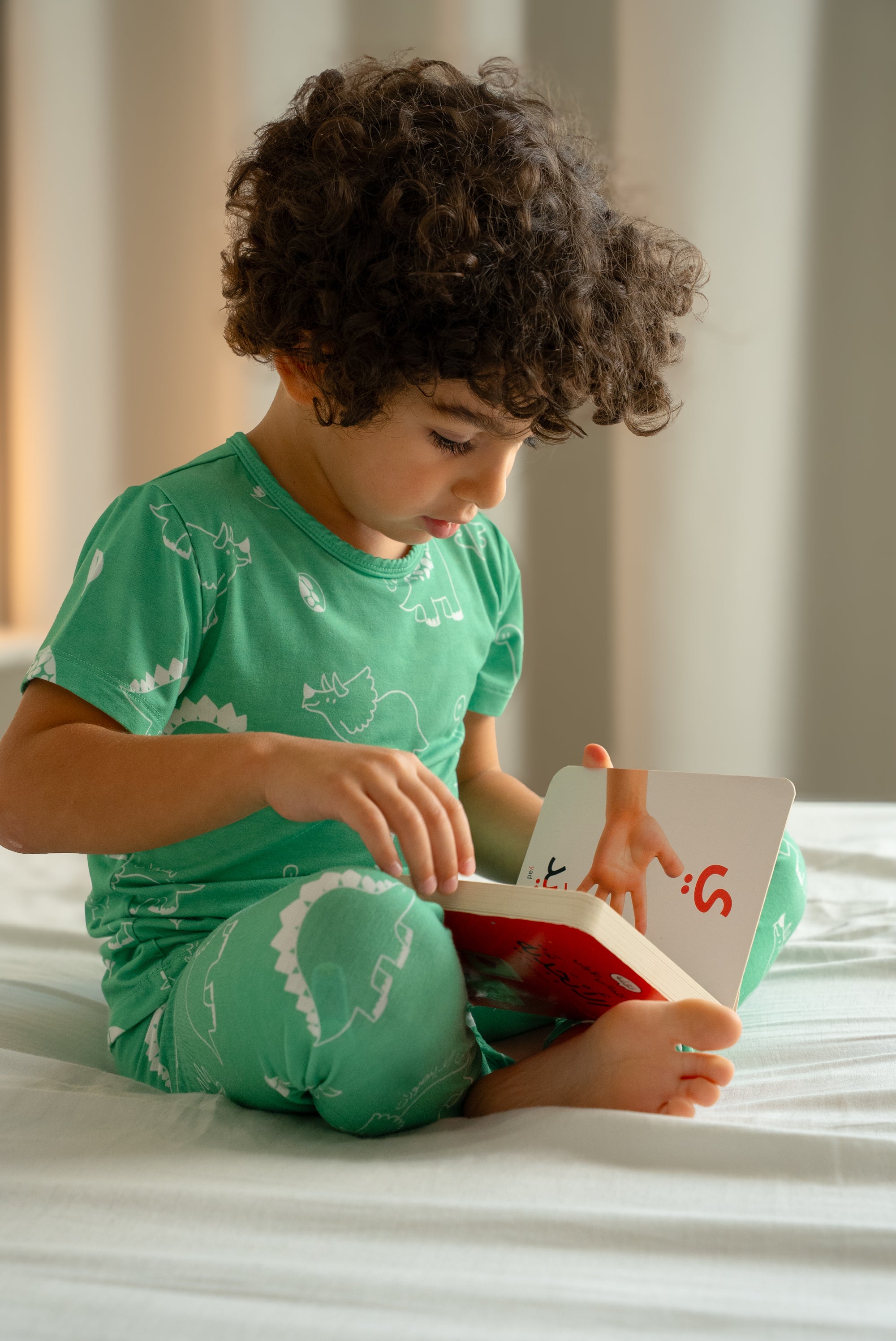 Child wearing our Dinosaur Pyjama, sitting on a bed with legs crossed and focused on reading a picture book.