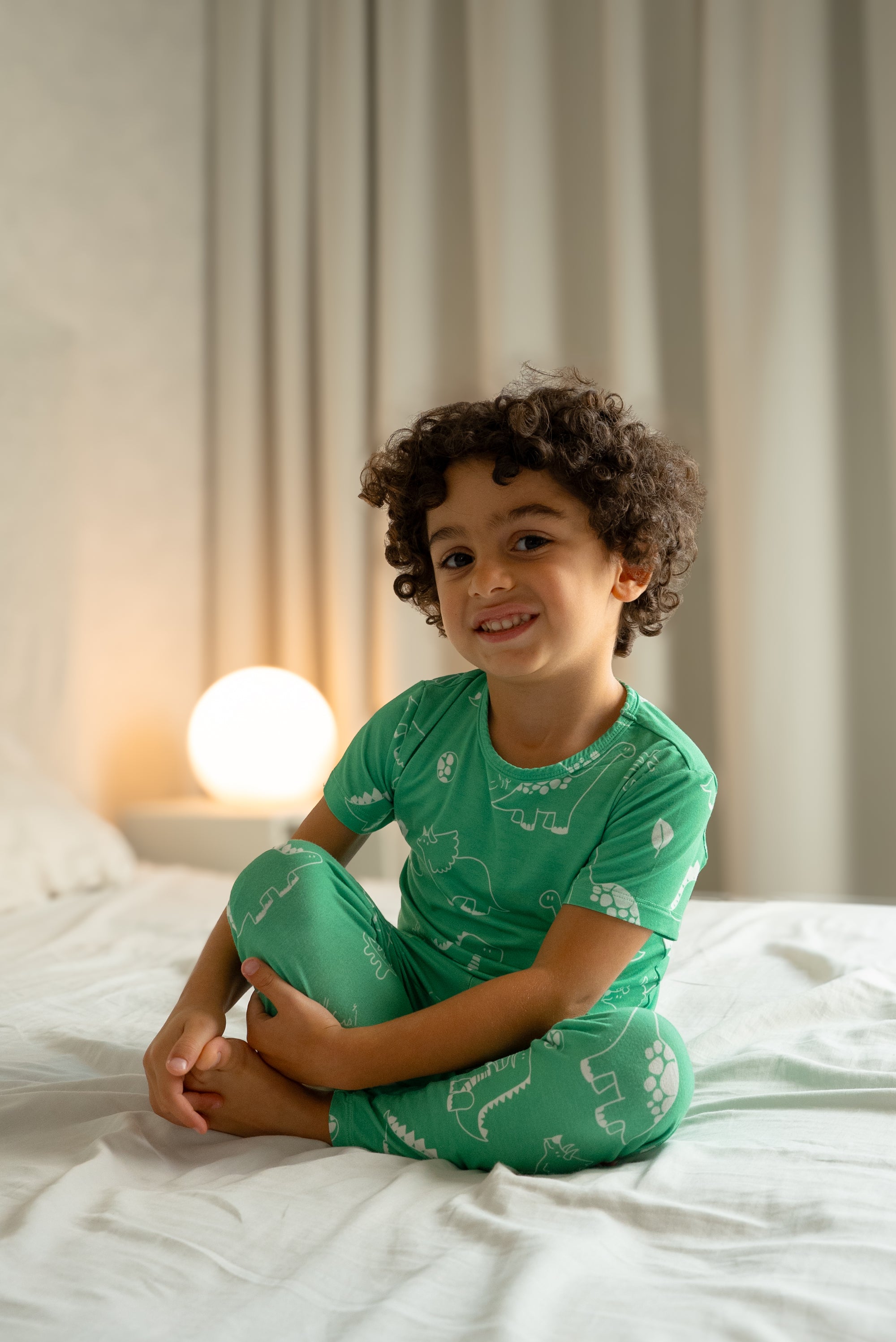 Child wearing our Dinosaur Pyjama, sitting comfortably on a bed with a gentle smile, showing the fun white dinosaur prints on soft green fabric.