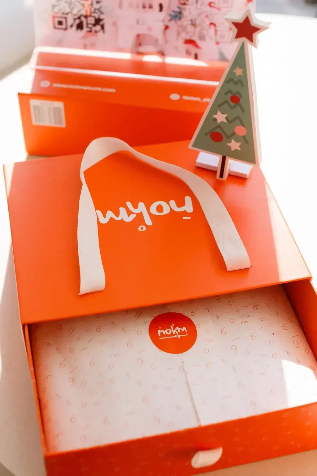 A festive orange Nohm gift box wrapped with care, featuring the Nohm Luxury Bedding Gift Set ready for gifting beside a small holiday tree.