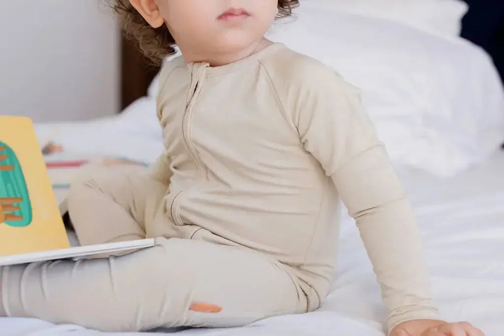 A baby sitting on a bed wearing the **Ramly Pyjama** from the **Grow with Nohm: Gift Set**, a beige bamboo one-piece featuring a front zipper for easy changes and a snug, comfy fit.

