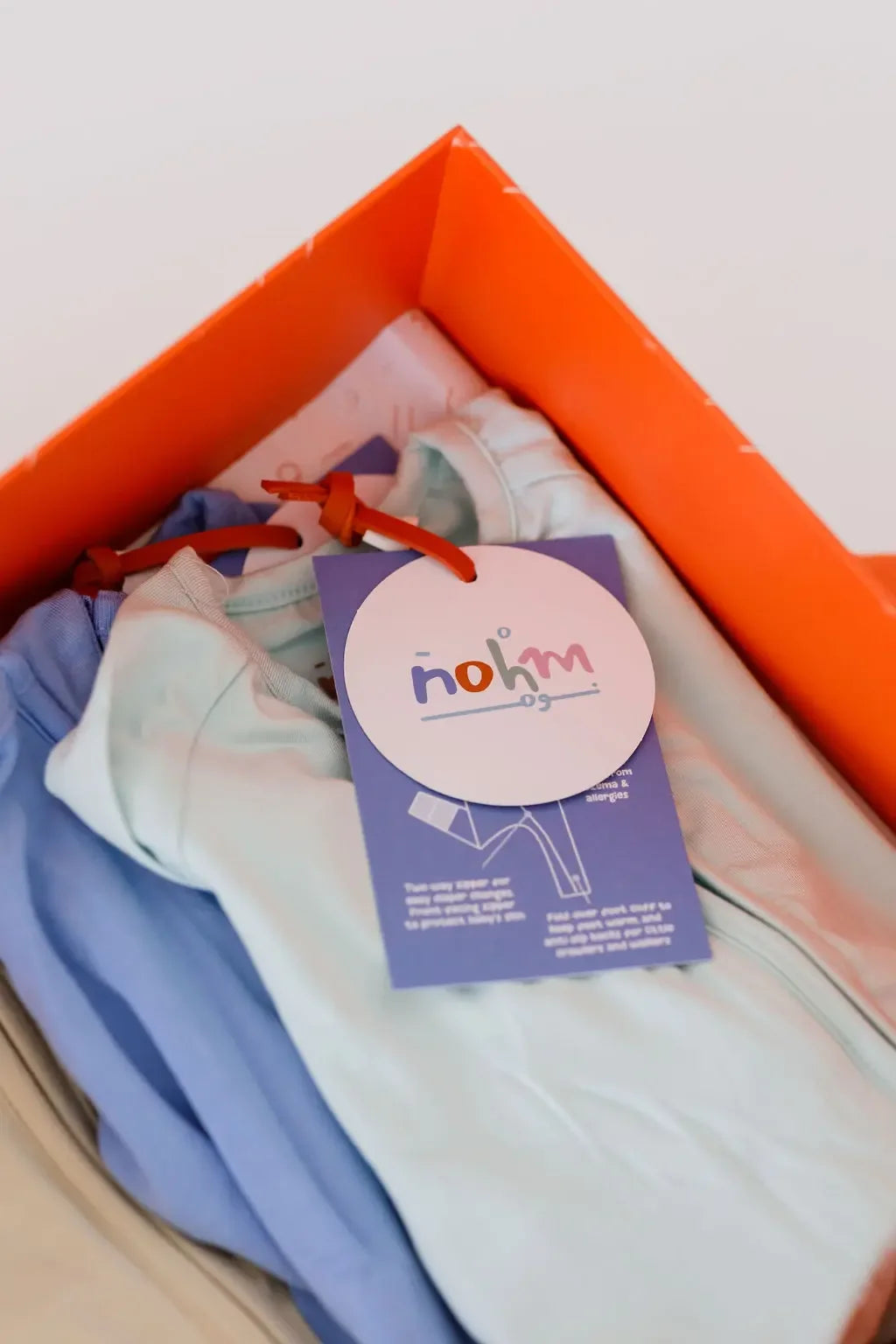 A close-up of the **Grow with Nohm: Gift Set**, showing neatly folded pastel baby pyjamas in an orange gift box with a branded Nohm tag.
