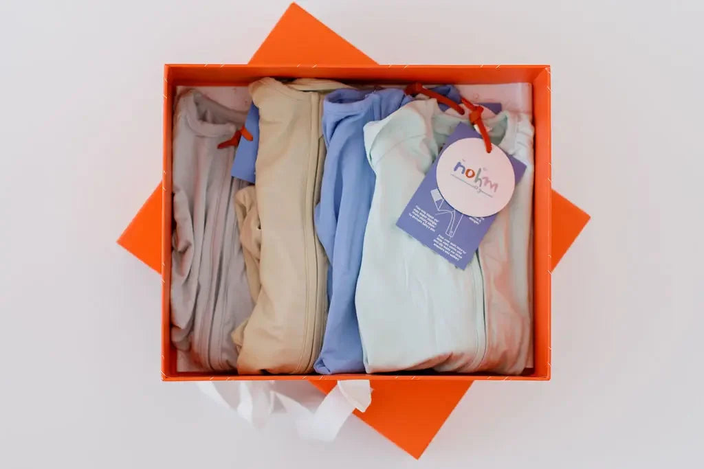 The **Grow with Nohm: Gift Set** displayed in an orange box, featuring four neatly folded baby pyjamas in soft neutral and pastel shades, each with a zipper design and Nohm tag.