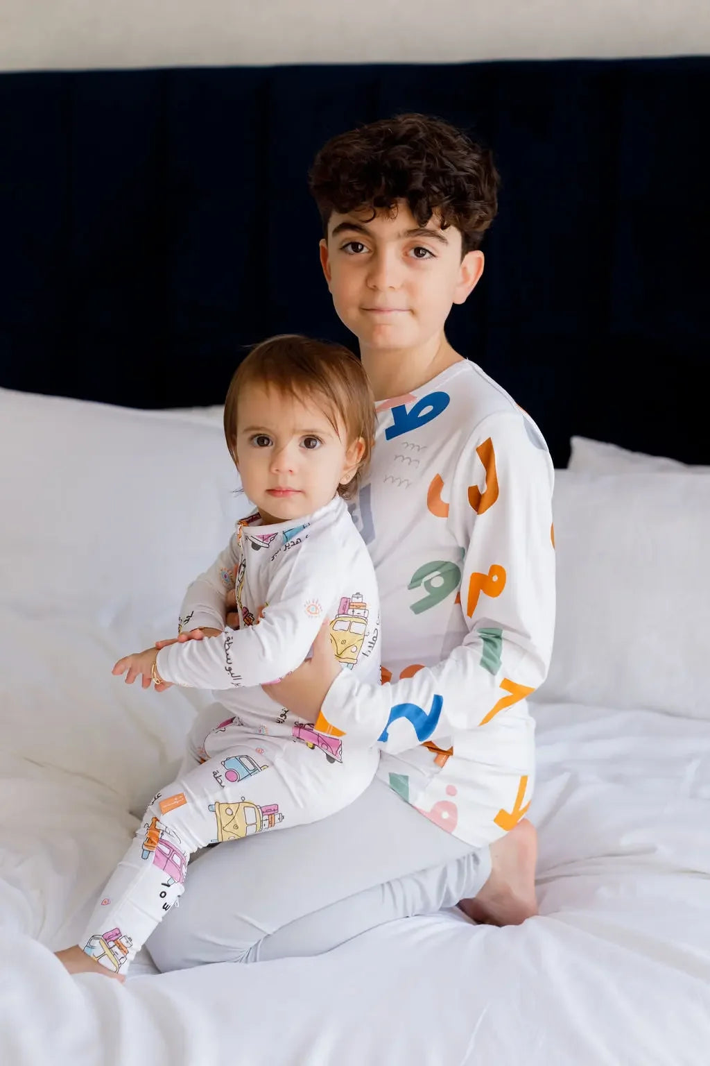 Two siblings sitting together on a bed, wearing our matching Huroof Long Sleeve Kids Pyjamas, celebrating comfort, connection, and Arabic-inspired design.