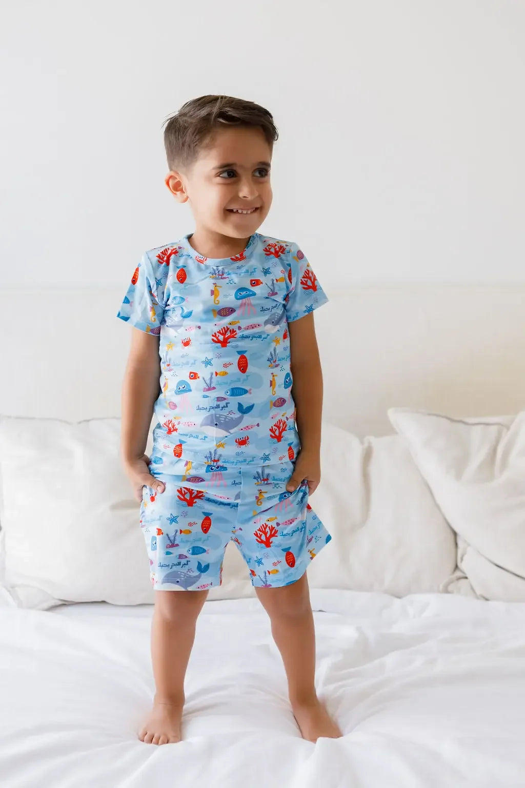 A happy boy standing on the bed while wearing our Kebr Al Baher short-sleeve set, featuring colorful sea creatures inspired by Fayrouz’s iconic song.