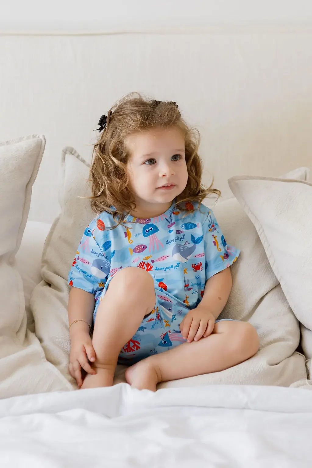 A sweet girl sitting comfortably on the bed while wearing our Kebr Al Baher short-sleeve set, bringing the ocean’s colors and charm to life.