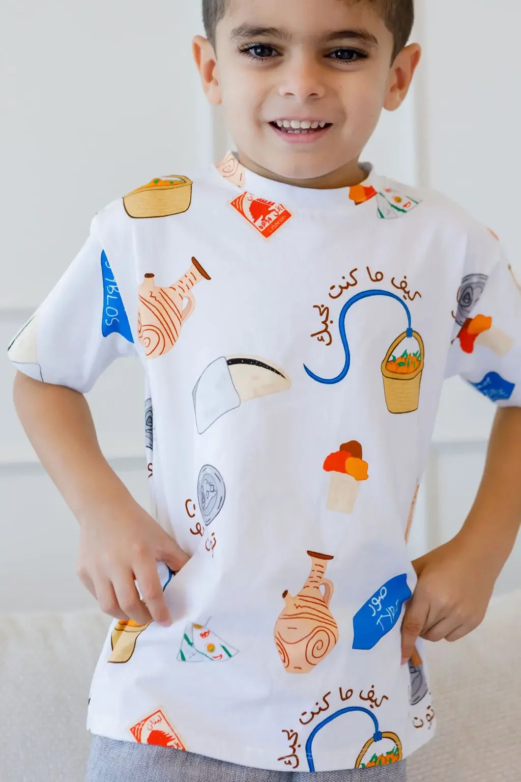 A smiling boy showing off our Lebanon T-Shirt, featuring playful prints of Lebanese icons like Man2ouche and Bonjus.