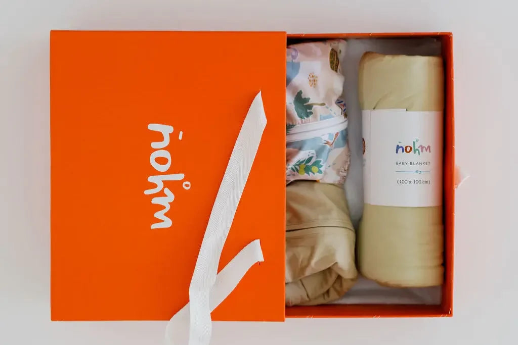 The **Neutral Nohm Gift Box – Newborn**, presented in a signature orange box, featuring a rolled beige Ramly blanket and two matching bamboo baby pyjamas, including the Bilady patterned design.
