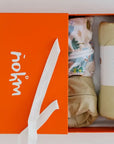 The **Neutral Nohm Gift Box – Newborn**, presented in a signature orange box, featuring a rolled beige Ramly blanket and two matching bamboo baby pyjamas, including the Bilady patterned design.
