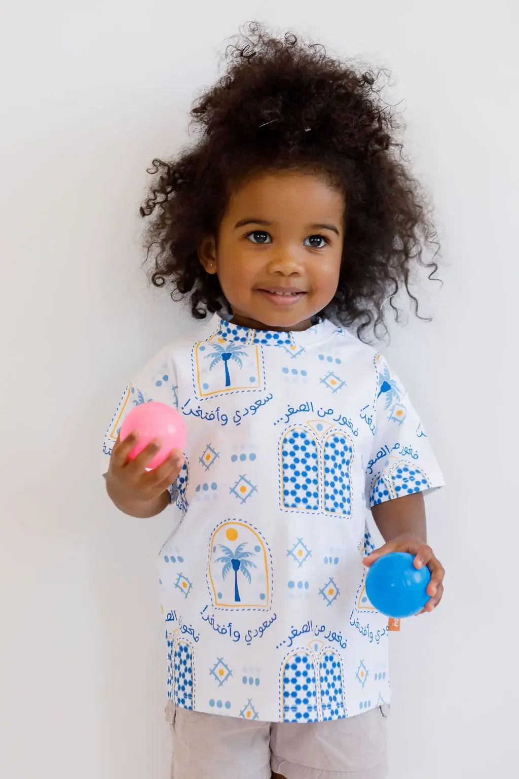 A happy girl holding colorful balls while wearing our Saudi T-Shirt, proudly showcasing the Kingdom’s cultural symbols and heritage.