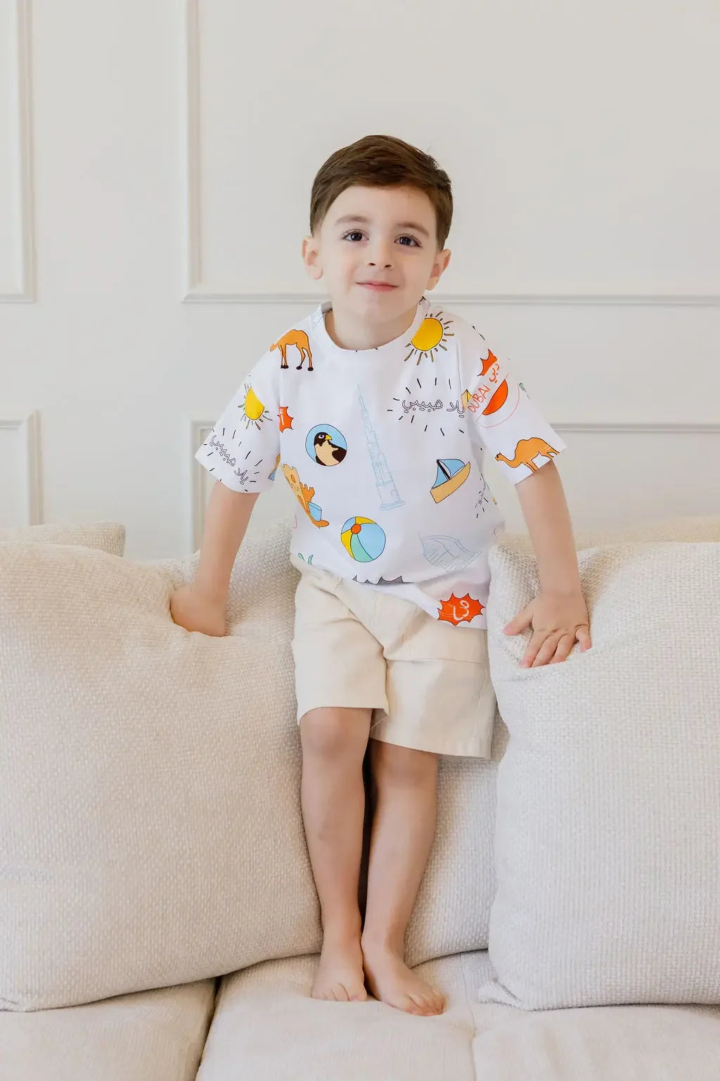 A cheerful boy standing on the couch wearing our UAE T-Shirt, showing off its fun and colorful heritage-inspired design.
