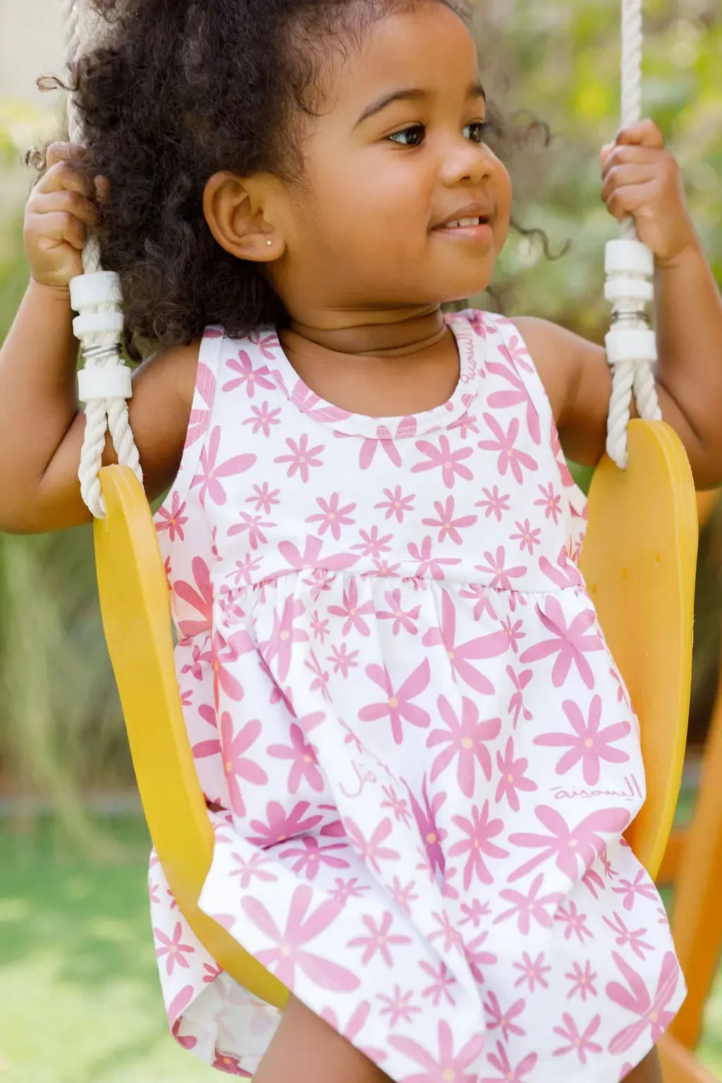 A joyful girl swinging outdoors while wearing our Yasmine Dress, blooming with pink jasmine flowers that capture playful elegance.